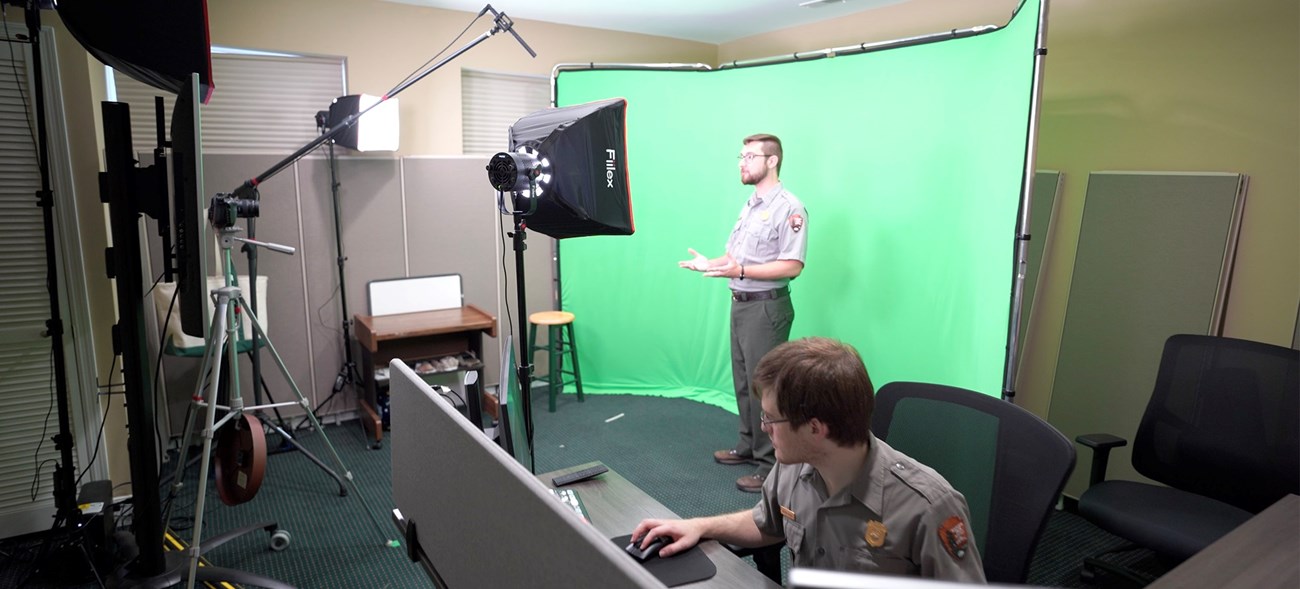 A behind the scenes look of a studio set up with a green screen. One ranger stands in front of the green screen while another runs a soundboard.