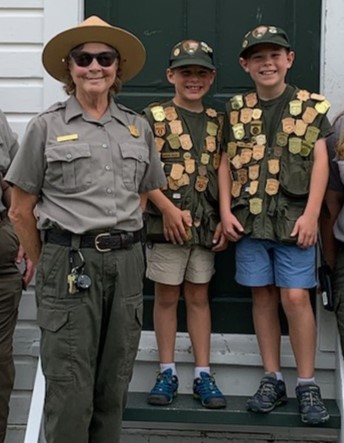 A park ranger standing beside two junior rangers displaying many junior ranger badges on their vests.