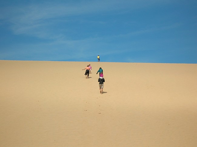 Kids run up the vast sand dune toward a deep blue sky