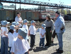 A Park Ranger and steam locomotive engineer talk with young school children.  The children are dressed in white shirts and wear engineer hats.