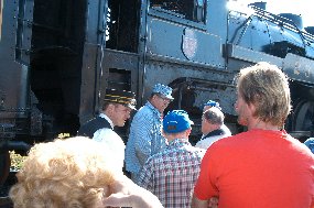 A conductor and engineer, both wearing historic costumes, talk with visitors next to a steam locomotive.