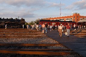 visitors walk accross railroad tracks with a steam locomotive and red passenger car sitting by a loading platform in the background