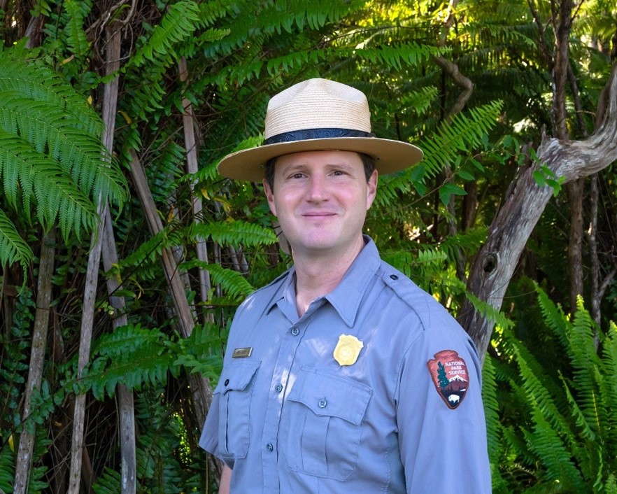 A man wears a National Park Service gray shirt and flat hat in front of a stand of ferns.