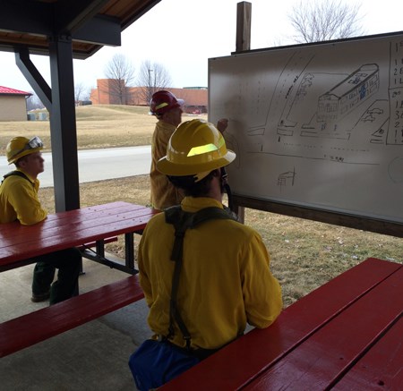 Three firefighters in yellow shirts and helmets sit in a pavillion