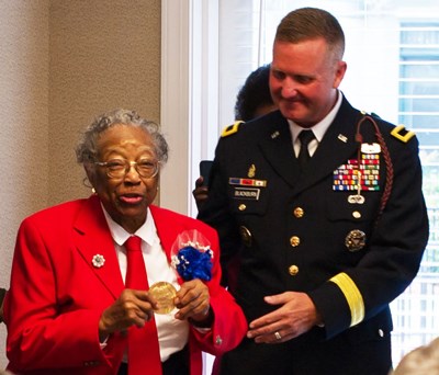 An older lady in a red coat holds up a gold medal while standing next to a man in a dark blue coat
