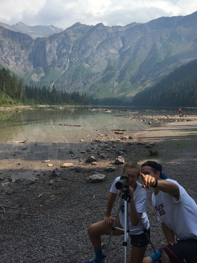 two people use a spotting scope in an alpine lake setting