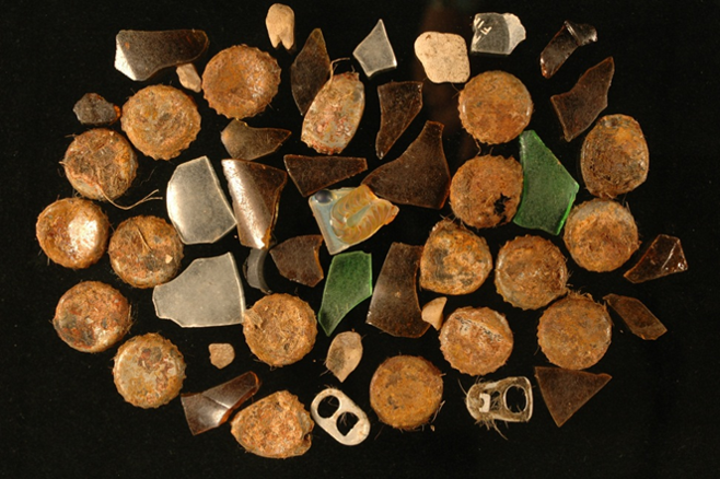 A pile of rusted bottle caps, pull tabs, and pieces of broken glass laid flat on a black background.
