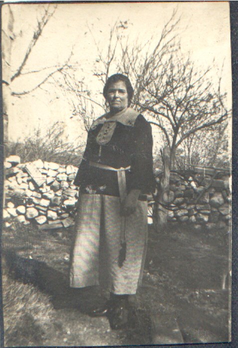 A woman wearing a long skirt, stands in front of a low stone wall with leafless trees on the other side.