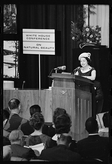 A seated audience watches a woman who speaks from behind a tall podium with two microphones, next to a sign that says "White House Conference on Natural Beauty"
