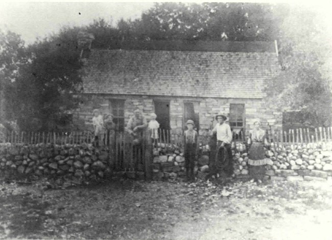 A group of seven people of varying ages stand along a stone and wood fence in front of a stone house.