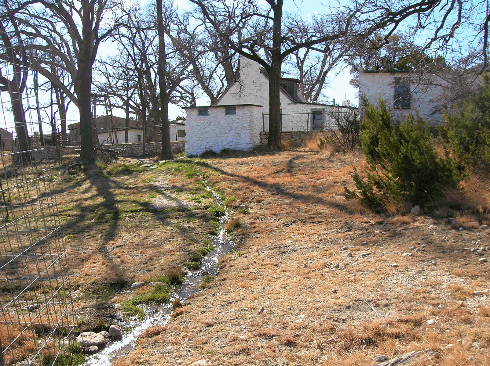 Water flows down a slope through an irrigation channel , looking toward the Ranch House cluster and leafless trees.