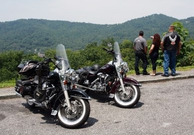 Three visitors looking at a mountain range standing next to two motorcycles after pulling over to an overlook area.