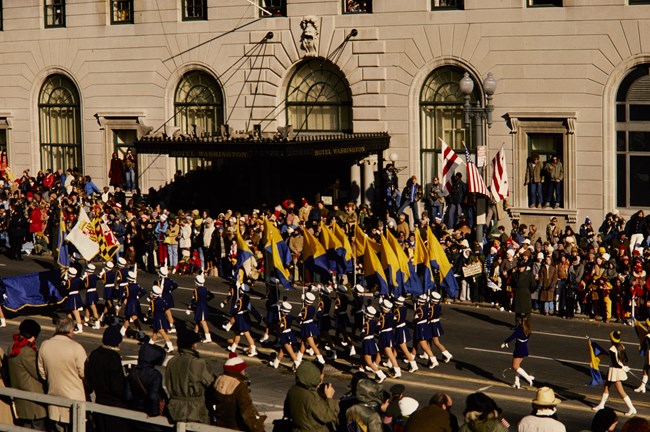A color guard walks carrying flags down a city street lined with observers