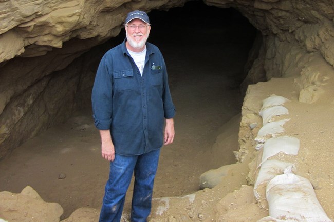 Man in blue shirt and jeans standing in front of cave.