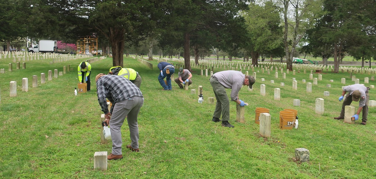 A group of seven people use spray bottles, brushes, and buckets to clean the surface of grave markers in a national cemetery