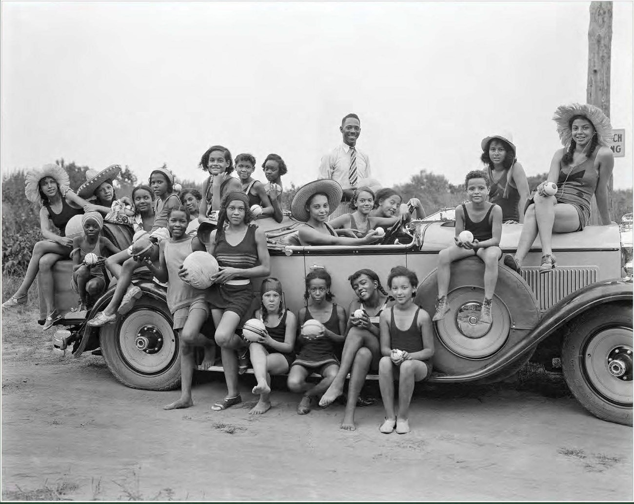 A group largely composed of young women playing ball at the beach pose by a vintage automobile.