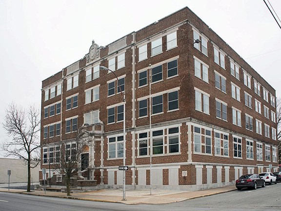 A four-story brick building with large rectangular windows on a street corner, featuring decorative stonework around the entrance and a small staircase leading up from the sidewalk.