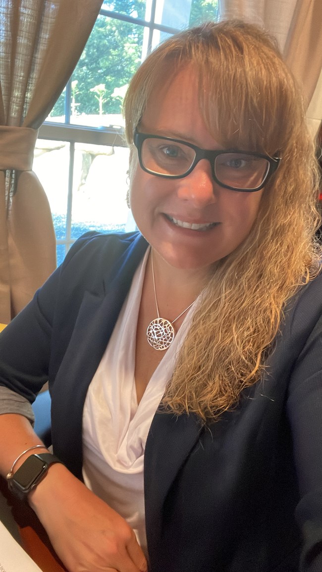 a woman with glasses and blond wavy hair smiles for the camera. She is wearing a dark colored blazer and white shirt.