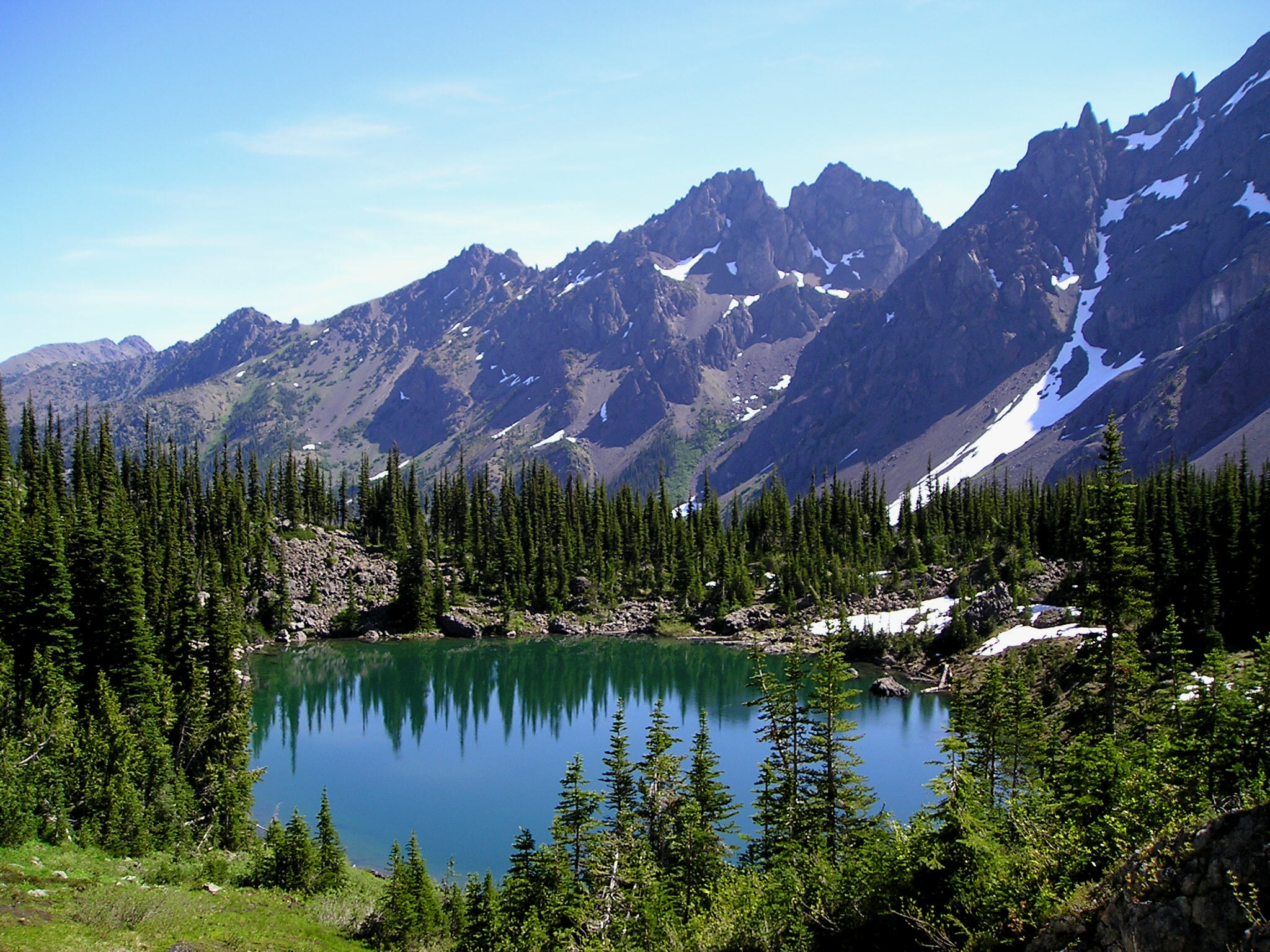Pointed conifers reflect in a still lake, surrounded by high, rocky mountains.
