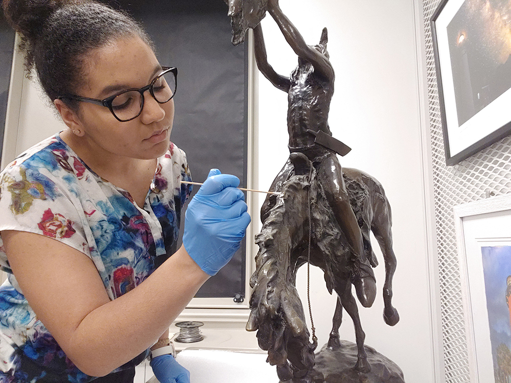 Woman with brown skin and hair wearing glasses and gloves is restoring a sculpture