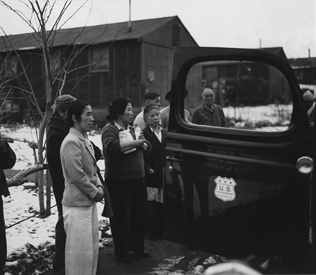 Group of women standing, facing right, near the open door of an automobile, buildings in the background.