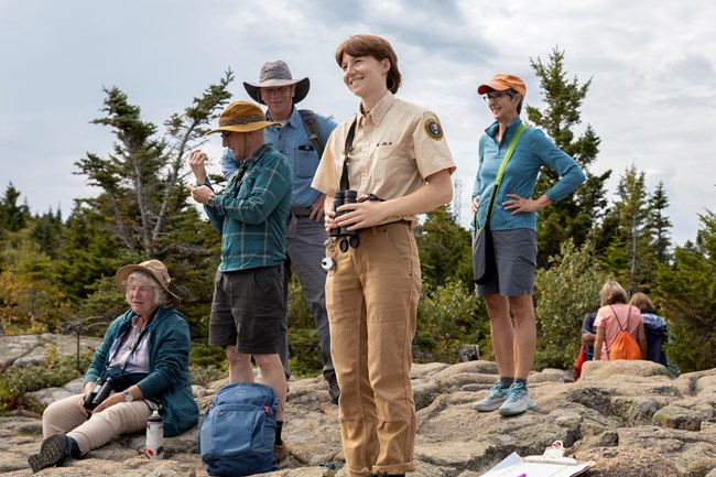 A smiling SCA Raptor Intern Holding a Pair of Binoculars During a Hawk Watch on Cadillac Mountain standing around four participating visitors.