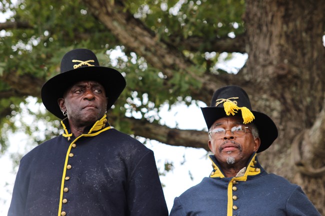 Two men dressed in blue uniforms with gold buttons stand in front of a tree