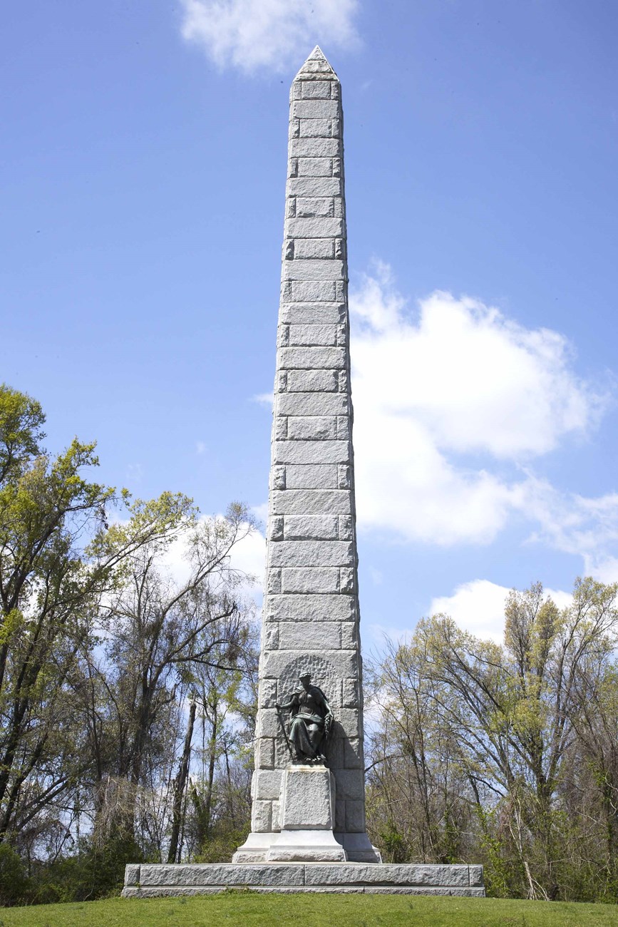 A tall white granite obelisk with bronze plaques on each side and in the center is bronze figure.