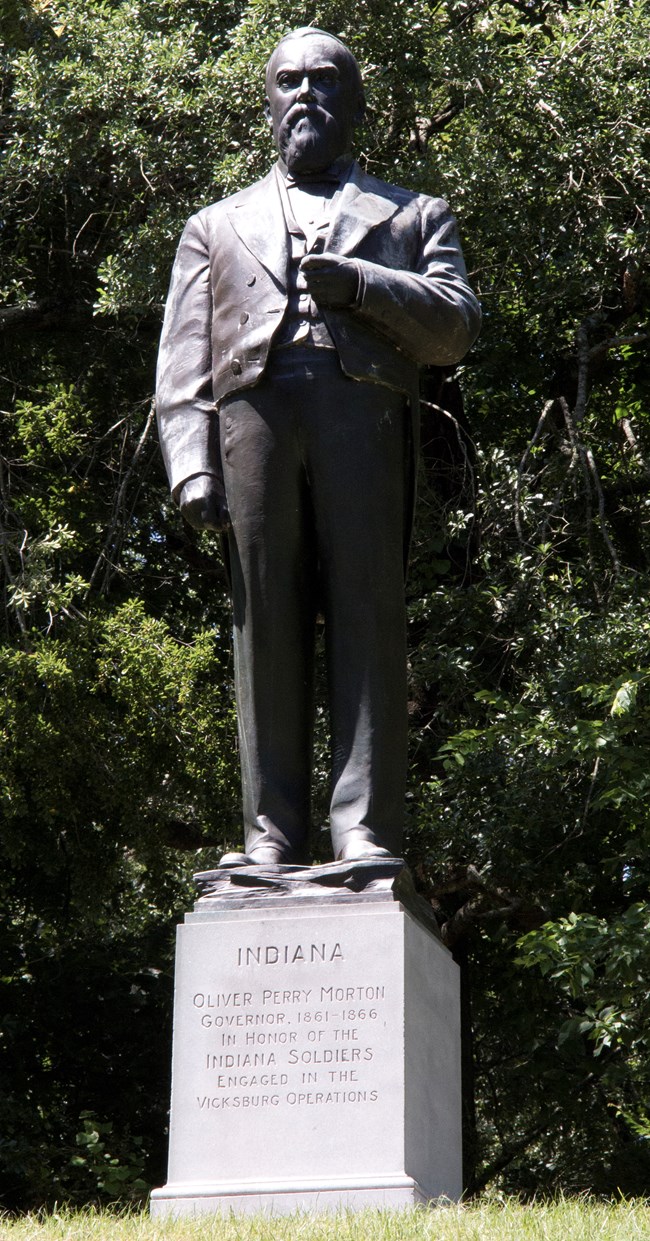 A bronze statue of a man in a formal suit. A pedestal sits below it with a short inscription