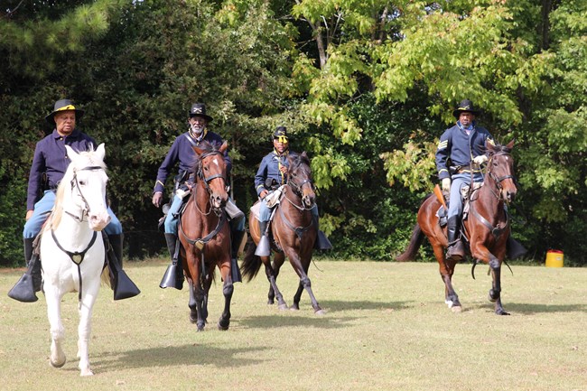 Four men dressed in blue uniforms with gold buttons ride horse through a field