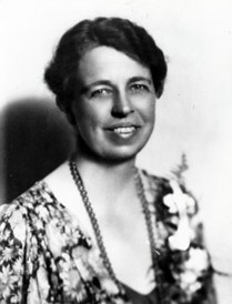 Eleanor Roosevelt poses for a portrait wearing a floral print shirt, a large arrangement of flowers pinned on her left shoulder, and a long string of beads.