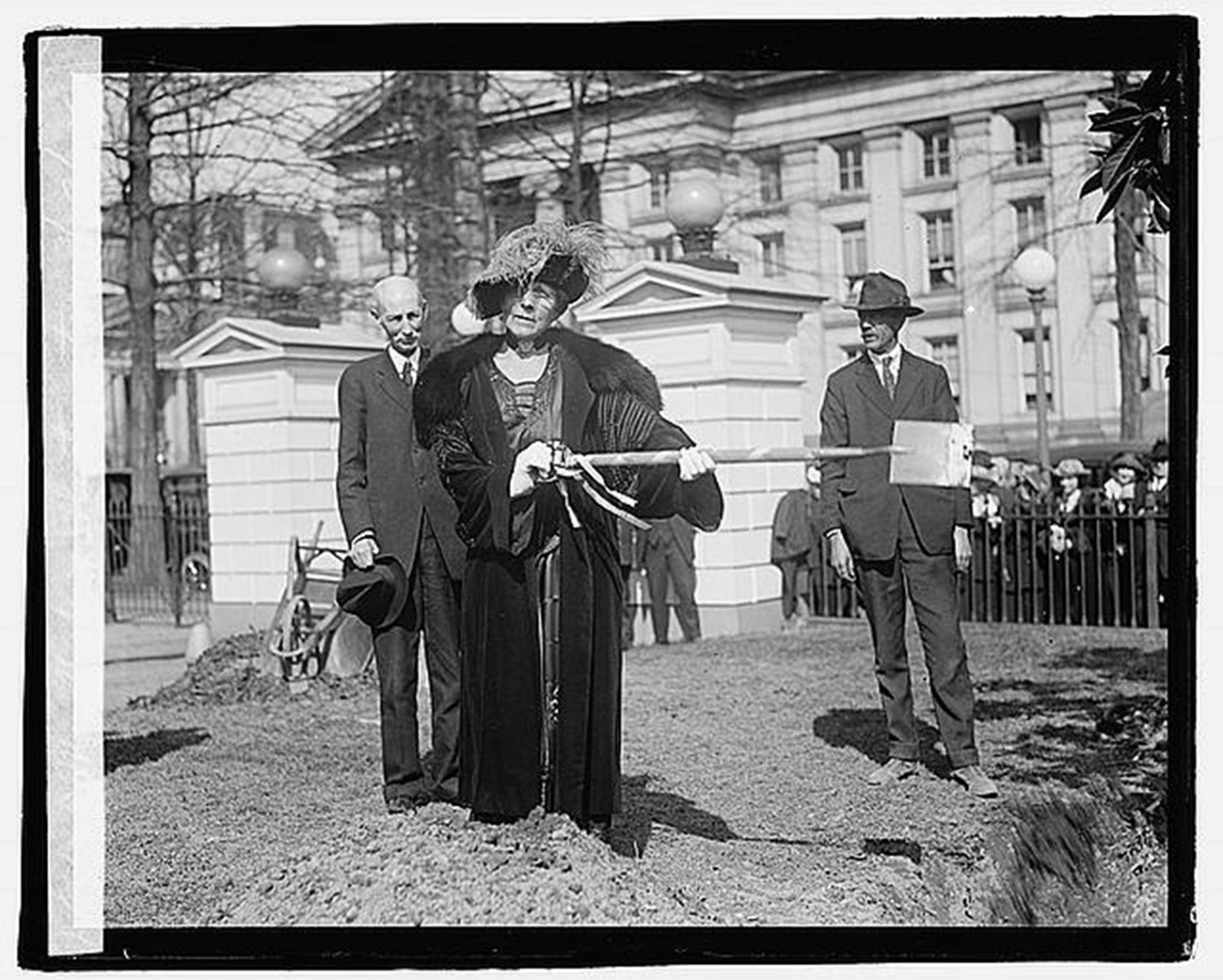 Florence Harding, in a fur coat and large, ornate hat, poses with a shovel in front of the Treasury Building near a White House gate.
