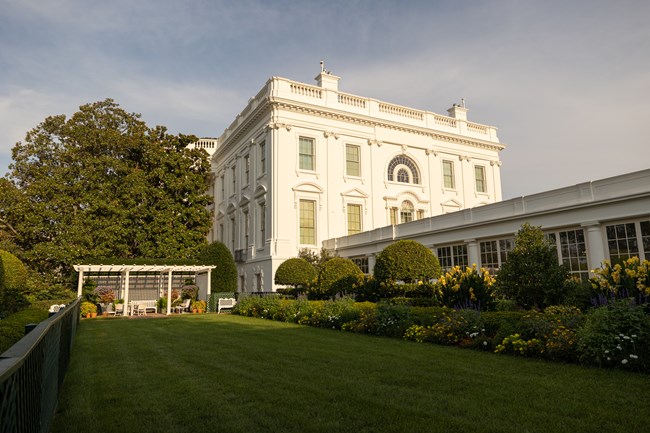A rectangular mowed lawn with a pergola at the rear and a garden of flowers by the White House.