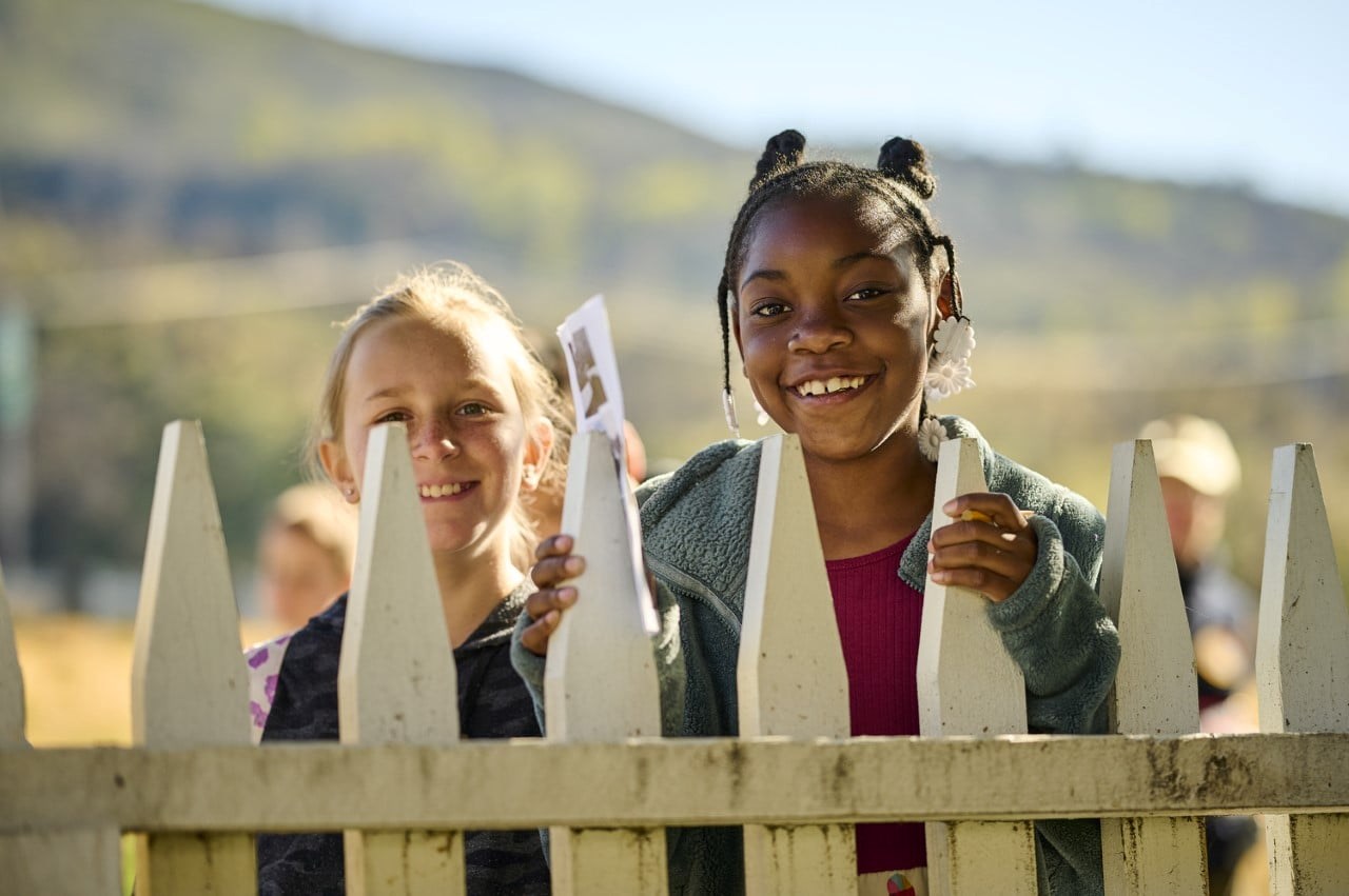 Two girls, one white and one African American, smiling with hands on a white fence.