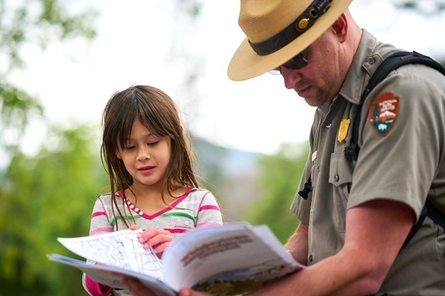 Photo of male park ranger wearing grey shirt, green pants, and flat hat, with girl with long brown hair. Ranger is holding a booklet