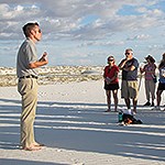 A man stands in the dunes barefoot addressing a group of people.