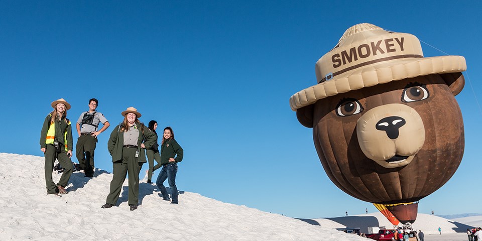 A group of employees stand on a sand dune with a giant Smokey Bear hot air balloon in the background.