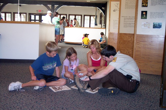A young male volunteer in a tan shirt and brown pants sits on the floor of the visitor center with a mother and two children helping them complete their junior ranger books.