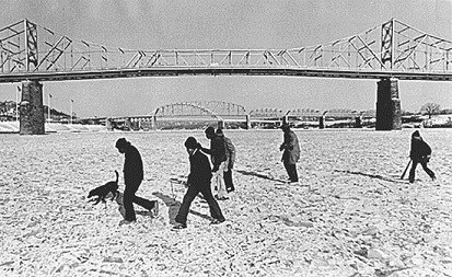 Several people walking across an icy surface with a suspension bridge in the background.