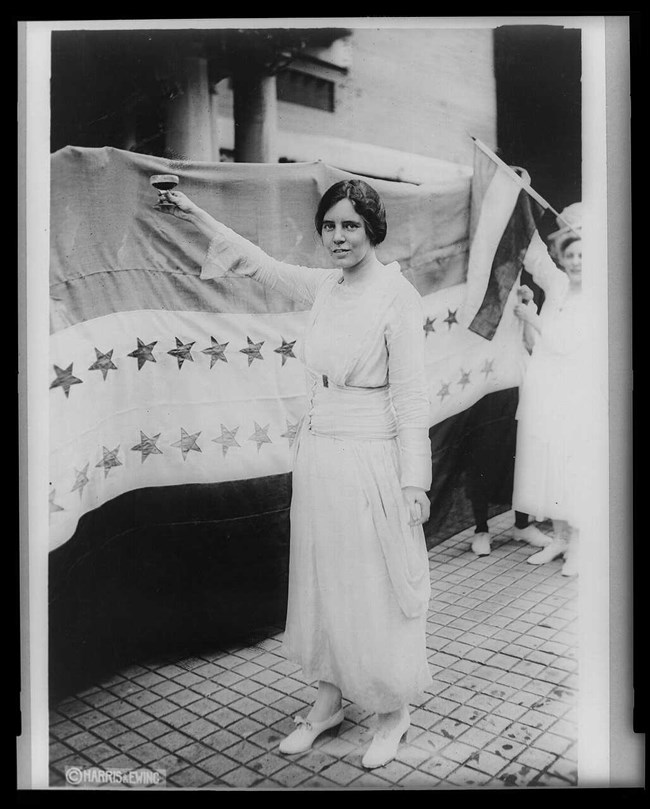 Alice Paul standing next to the Suffrage Flag holding a glass in a toast salute.