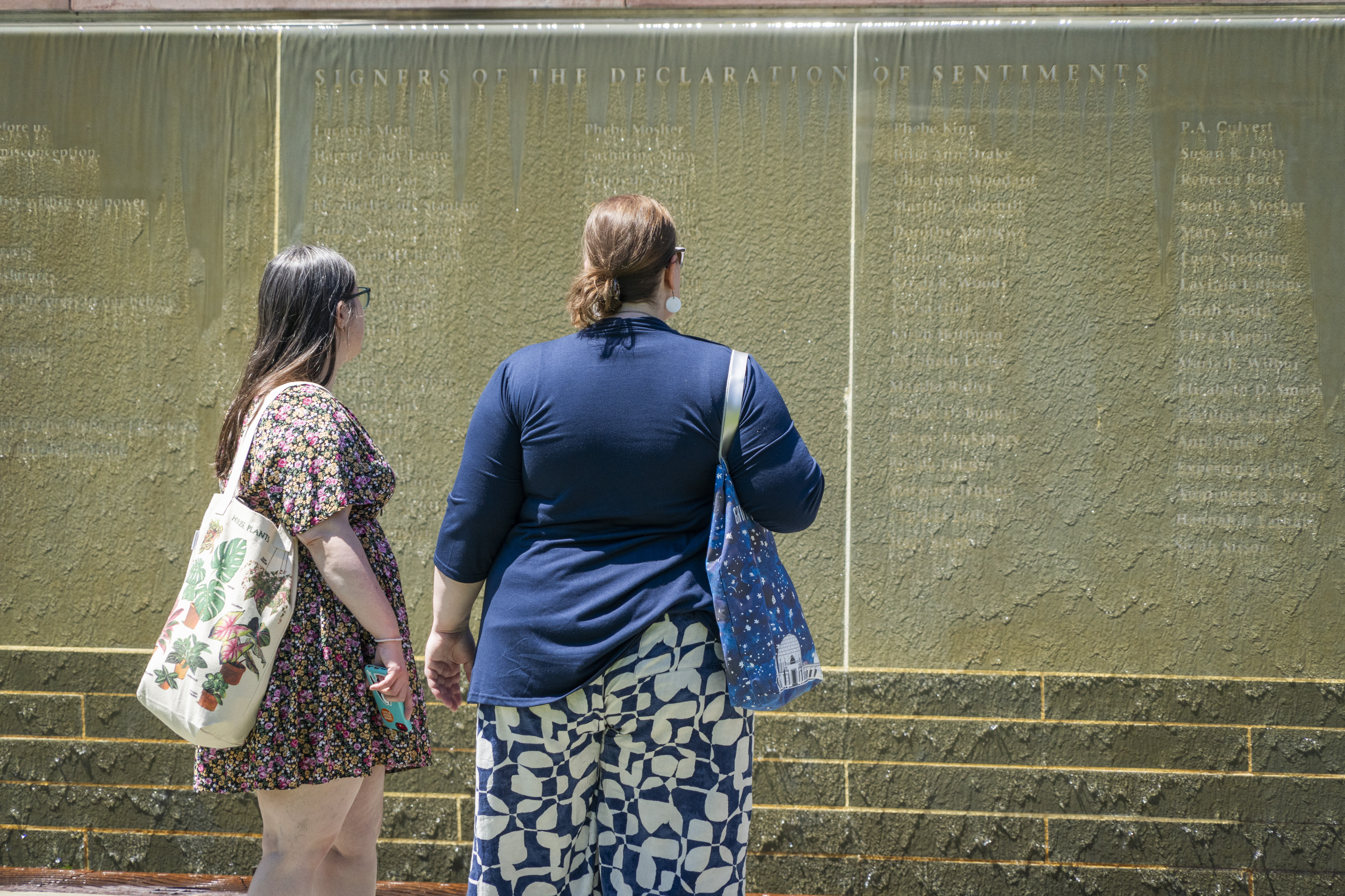 Two women stand and read the names of the signers of the Declaration of Sentiments inscribed on a stone wall; water flows down the wall and runs over the names.