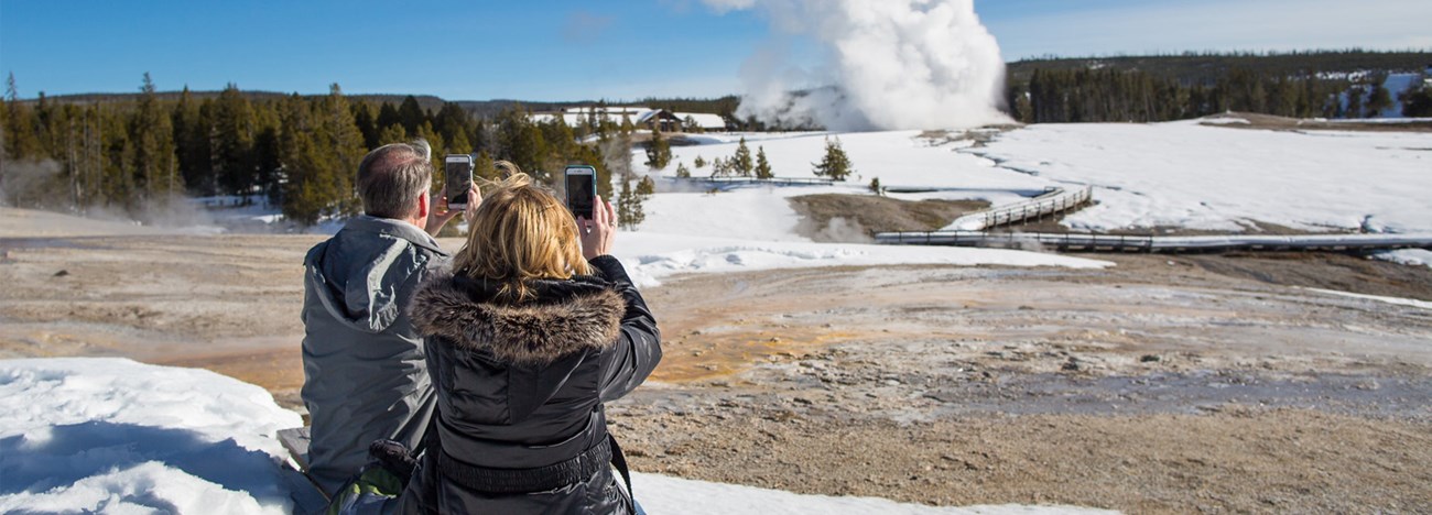 A man and a woman sit on a bench and hold their phones up to film a geyser erupting in the background.