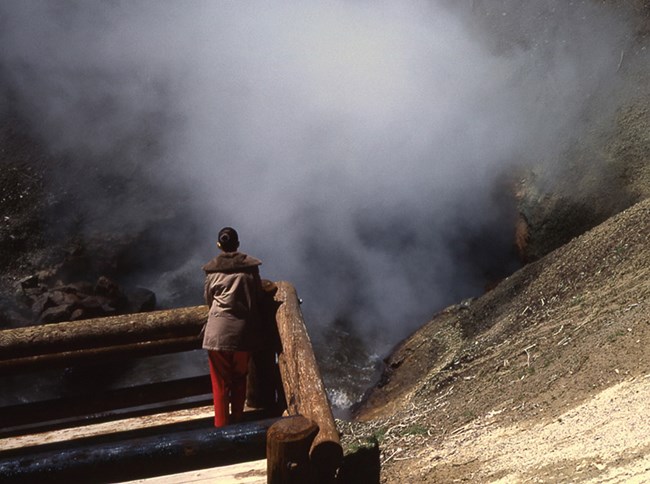 Visitor standing at a boardwalk rail overlooking billowing steam from Dragon's Mouth hot spring