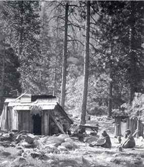 Two American Indians sit on ground by their simple housing