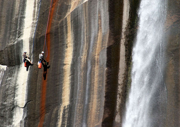 Two men hang from climbing ropes against a lichen-covered vertical rock face