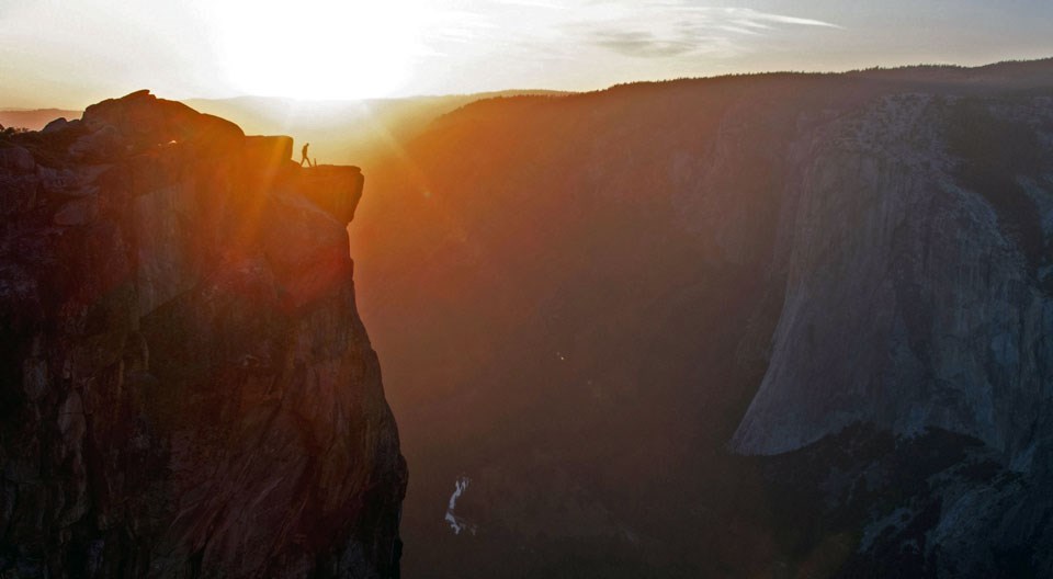 Person standing near the edge of a cliff, watching the sun set.