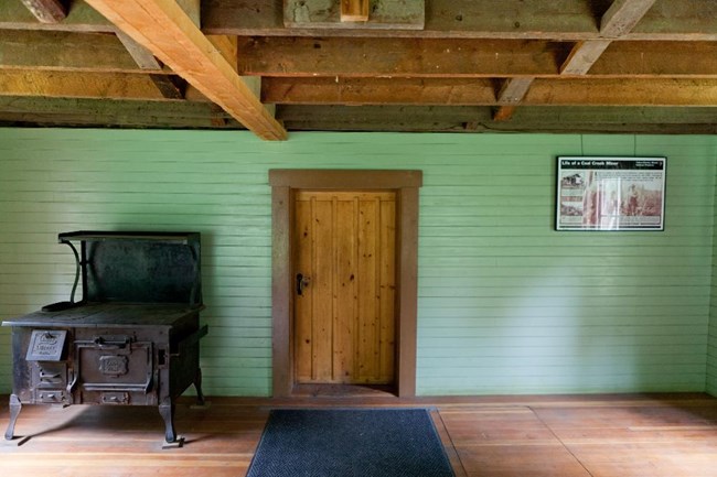 View of wooden door, set into a green painted wall. An metal wood burning stove is to the left of the door.