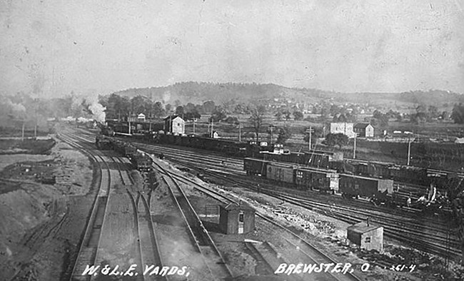 Wheeling and Lake Erie Rail Yard, Brewster, Ohio. Postcard, ca. 1910. Wikimedia. Black and white photo postcard overlooking a busy rail yard. There are several tracks, switches, and small buildings. Lines of rail cars sit on the tracks. In the distance are low mountains.