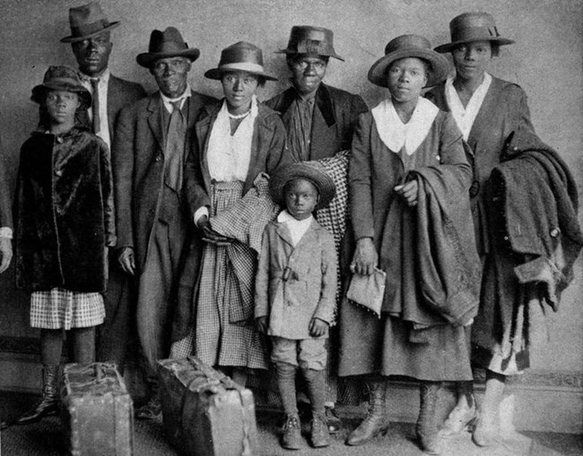 The Arthur family arrives in Chicago on August 30, 1920. Two months earlier, a mob burned two of their sons alive in a lynching in Paris, Texas. Originally published in The Chicago Defender, September 4, 1920. Black and white photo of a Black family (two men, five women, and a young boy) dressed in suits and dresses, all wearing hats. In front of them are two suitcases.