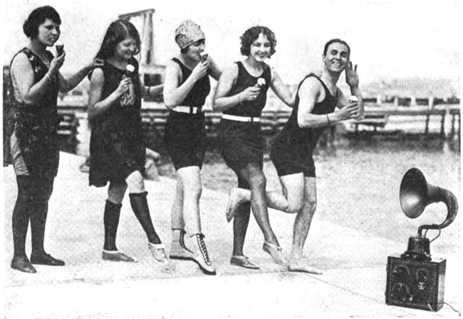 1920s actors eat ice cream and dance at the beach to the music of an early radio, August 1923. Wireless Age August 1923, p. 26. Black and white photo pf four white women and a white man wearing 1920s bathing suits and eating ice cream cones. A box with knobs and a trumpet-shaped amplifier (the radio) sits on the beach in front of them.